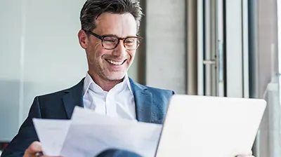 Man in business attire smiling while looking at laptop and holding documents.